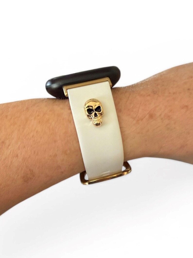 Op de afbeelding: Witte horlogeband met een gouden schedelbedel. De band heeft goudkleurige hardware en is bevestigd aan een zwarte wijzerplaat.