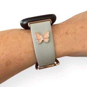 May include: A light gray watch band with a rose gold clasp and a pink butterfly charm. The butterfly is embellished with clear crystals. The watch face is black.