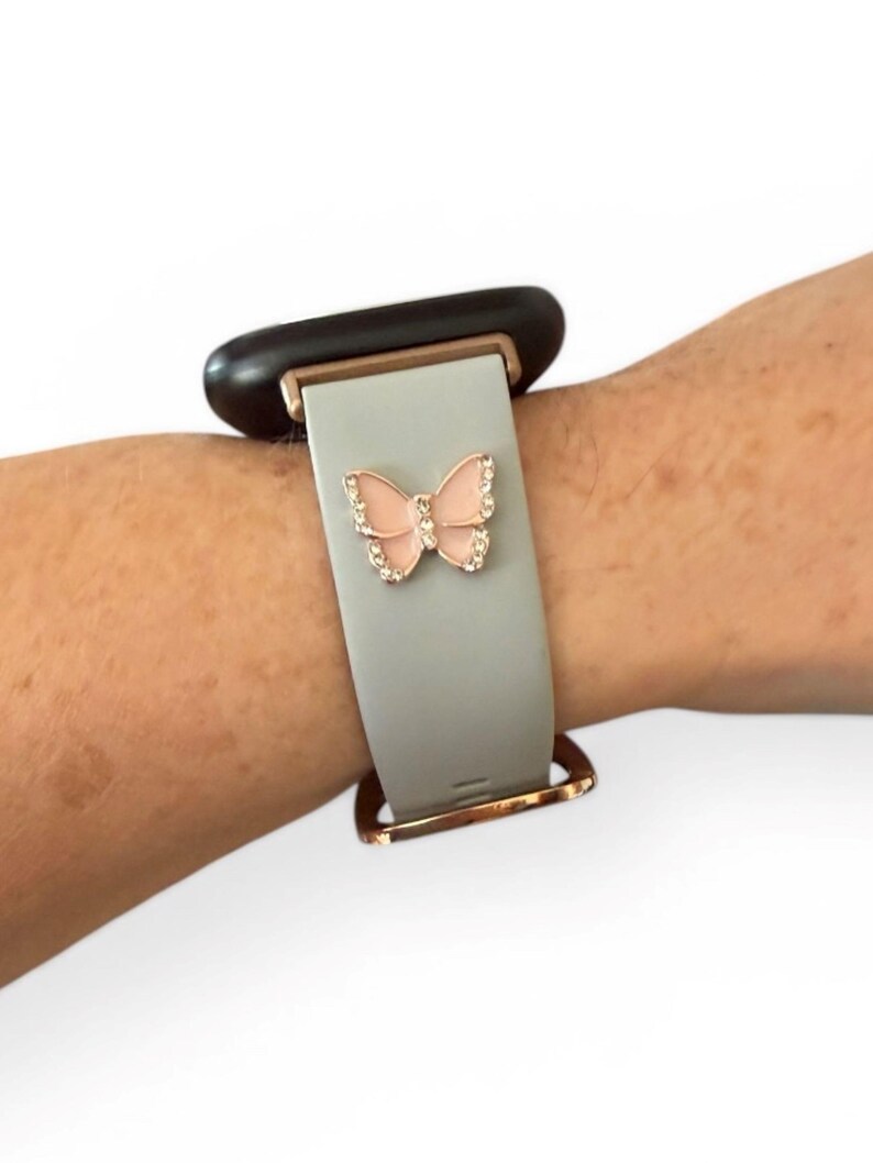 May include: A light gray watch band with a rose gold clasp and a pink butterfly charm. The butterfly is embellished with clear crystals. The watch face is black.