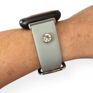 Peut inclure: Un bracelet de montre gris clair avec une boucle argent&eacute;e et un grand ornement rond en cristal transparent. Le cadran de la montre est noir. Le bracelet est attach&eacute; &agrave; un poignet.