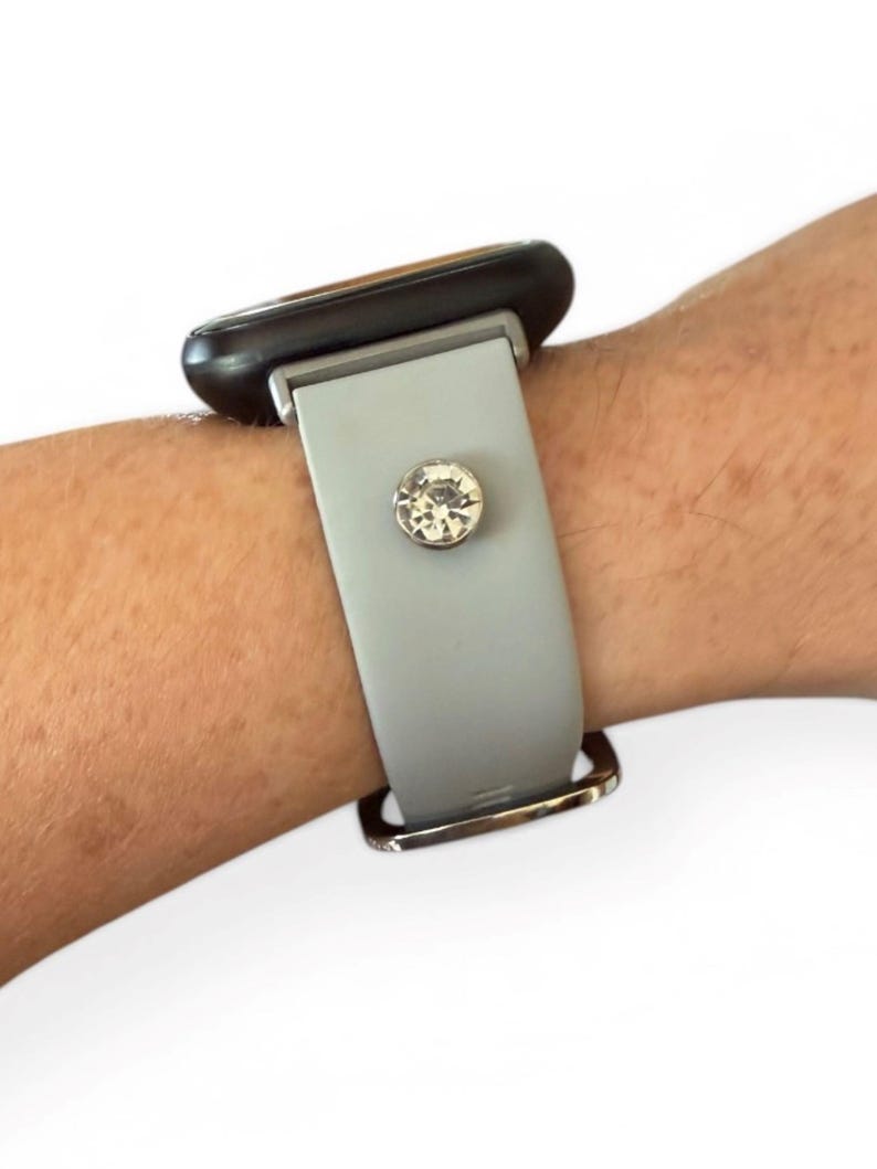 Peut inclure: Un bracelet de montre gris clair avec une boucle argent&eacute;e et un grand ornement rond en cristal transparent. Le cadran de la montre est noir. Le bracelet est attach&eacute; &agrave; un poignet.