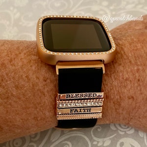 K&ouml;nnte beinhalten: Ros&eacute;goldene Smartwatch mit schwarzem Silikonarmband und einer mit Kristallen besetzten L&uuml;nette. Das Armband hat austauschbare Charms mit den Worten "Blessed" und "Faith" darauf.