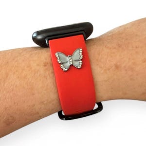 May include: Red smartwatch band with a silver butterfly charm. The butterfly is embellished with small, clear crystals. The watch case is black.