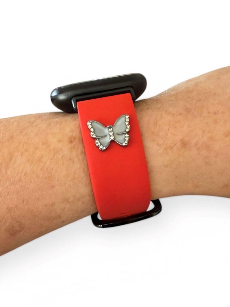May include: Red smartwatch band with a silver butterfly charm. The butterfly is embellished with small, clear crystals. The watch case is black.