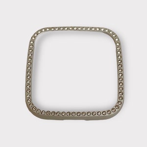 May include: A silver square-shaped watch band with a row of clear rhinestones around the edge.