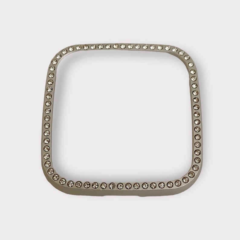 May include: A silver square-shaped watch band with a row of clear rhinestones around the edge.