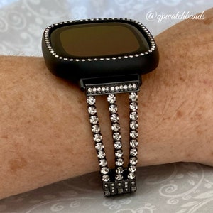 K&ouml;nnte beinhalten: Schwarze Smartwatch mit einem schwarzen Armband mit Strasssteinen. Das Armband hat drei Reihen mit Strasssteinen.
