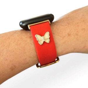 May include: Red watch band with a gold buckle and a cream-colored butterfly charm with rhinestone accents. The watch band is attached to a black and gold smartwatch.