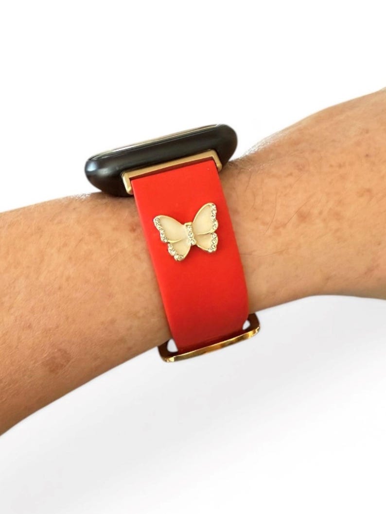 May include: Red watch band with a gold buckle and a cream-colored butterfly charm with rhinestone accents. The watch band is attached to a black and gold smartwatch.