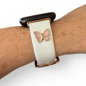 May include: White watch band with a rose gold buckle and a pink butterfly charm with clear rhinestones. The watch band is attached to a black watch face.
