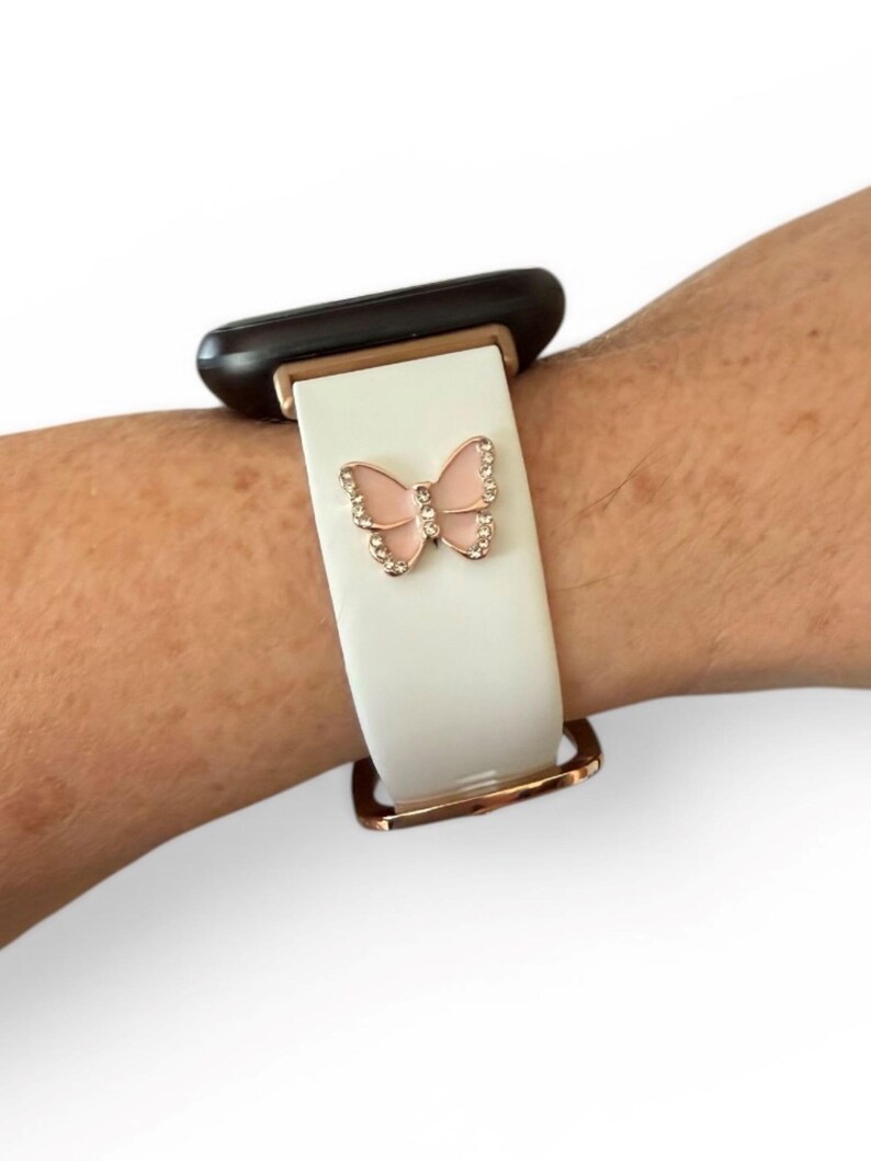 May include: White watch band with a rose gold buckle and a pink butterfly charm with clear rhinestones. The watch band is attached to a black watch face.