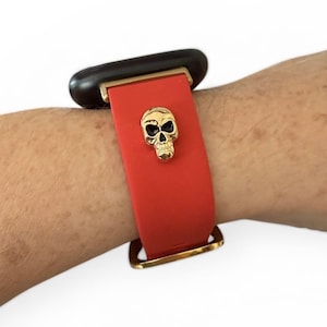 Op de afbeelding: Rode horlogeband met een gouden schedelbedel. De band is bevestigd met goudkleurige hardware. De wijzerplaat van het horloge is zwart.