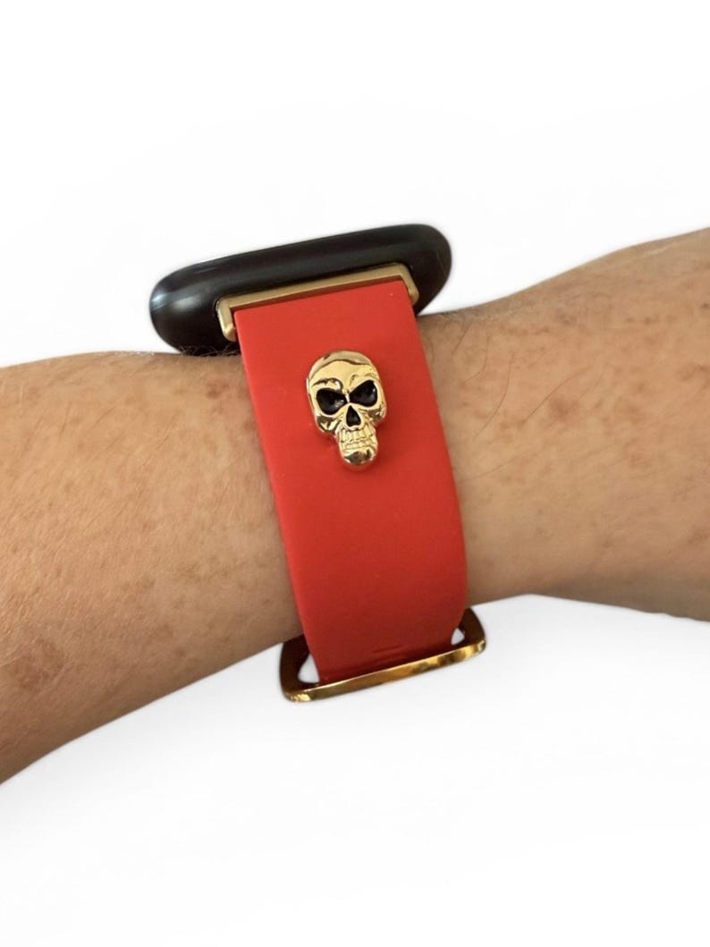 Op de afbeelding: Rode horlogeband met een gouden schedelbedel. De band is bevestigd met goudkleurige hardware. De wijzerplaat van het horloge is zwart.