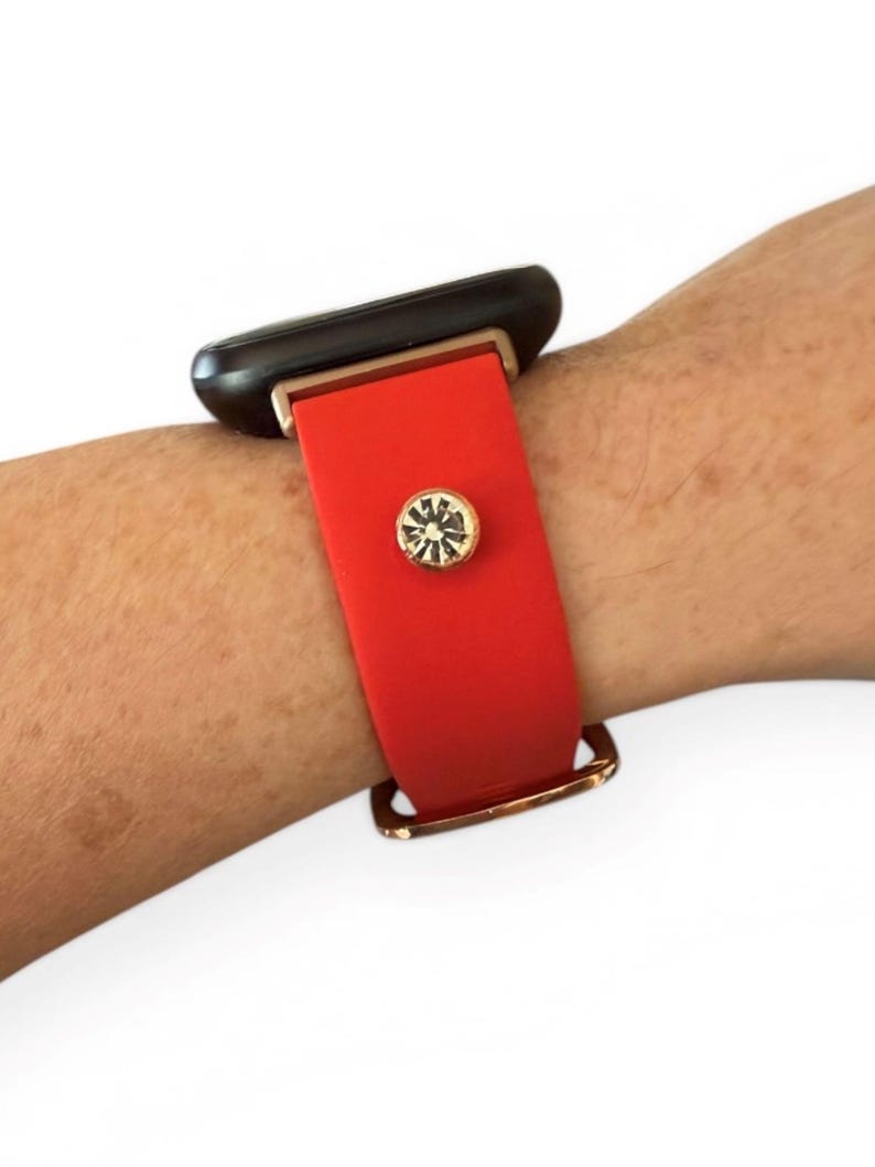 Peut inclure: Un bracelet de montre en silicone rouge avec des &eacute;l&eacute;ments dor&eacute;s et un accent d&eacute;coratif en cristal. Le bracelet est fix&eacute; &agrave; une montre intelligente noire.