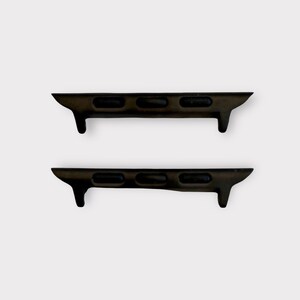 May include: Two dark brown watch band adapters. Each adapter has three rectangular holes and a small, downward-facing tab on one end. The adapters are designed to connect a watch band to a watch face.