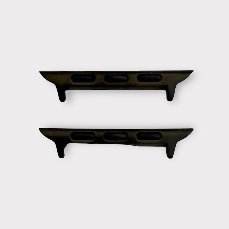 May include: Two dark brown watch band adapters. Each adapter has three rectangular holes and a small, downward-facing tab on one end. The adapters are designed to connect a watch band to a watch face.