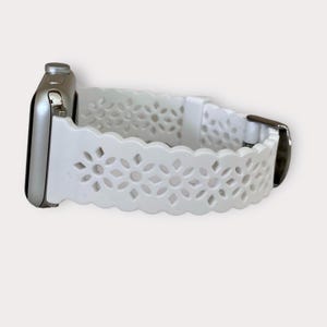May include: White silicone Apple Watch band with a delicate floral cutout design. The band has a scalloped edge and a silver buckle.