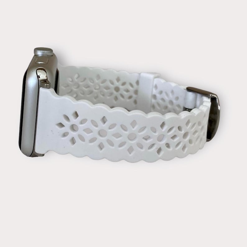 May include: White silicone Apple Watch band with a delicate floral cutout design. The band has a scalloped edge and a silver buckle.