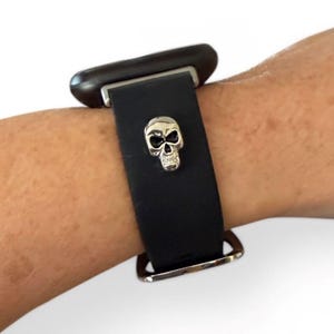 Op de afbeelding: Zwarte leren horlogeband met een zilveren schedelbedel. De band heeft een zilveren gesp en een donkerbruine wijzerplaat. De horloge zit om de pols van een persoon.