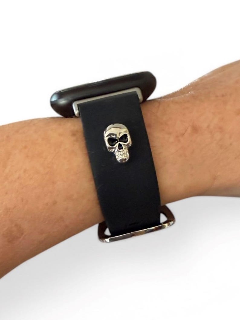 Op de afbeelding: Zwarte leren horlogeband met een zilveren schedelbedel. De band heeft een zilveren gesp en een donkerbruine wijzerplaat. De horloge zit om de pols van een persoon.