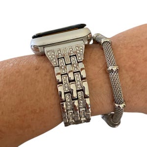 Puede incluir: Un reloj plateado con una correa con incrustaciones de pedrería y una pulsera de malla plateada. La correa del reloj está hecha de eslabones rectangulares con pedrería incrustada. La pulsera de malla está al lado del reloj.