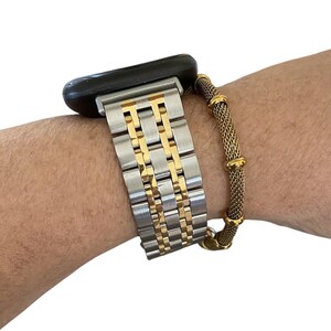 Peut inclure: Un bracelet de montre argent&eacute; et dor&eacute; est fix&eacute; &agrave; une montre intelligente noire. Un bracelet en maille dor&eacute;e est port&eacute; sur le poignet &agrave; c&ocirc;t&eacute; de la montre. Le bracelet de montre a une base argent&eacute;e avec des accents dor&eacute;s.