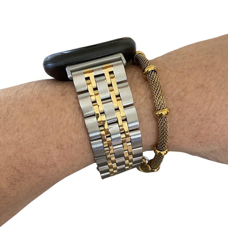 Peut inclure: Un bracelet de montre argent&eacute; et dor&eacute; est fix&eacute; &agrave; une montre intelligente noire. Un bracelet en maille dor&eacute;e est port&eacute; sur le poignet &agrave; c&ocirc;t&eacute; de la montre. Le bracelet de montre a une base argent&eacute;e avec des accents dor&eacute;s.