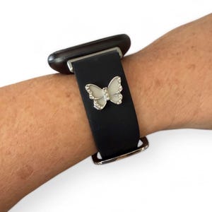 May include: Black watch band with a silver buckle and a decorative butterfly charm. The butterfly is white with silver-colored rhinestone accents. The watch band is attached to a black smartwatch.