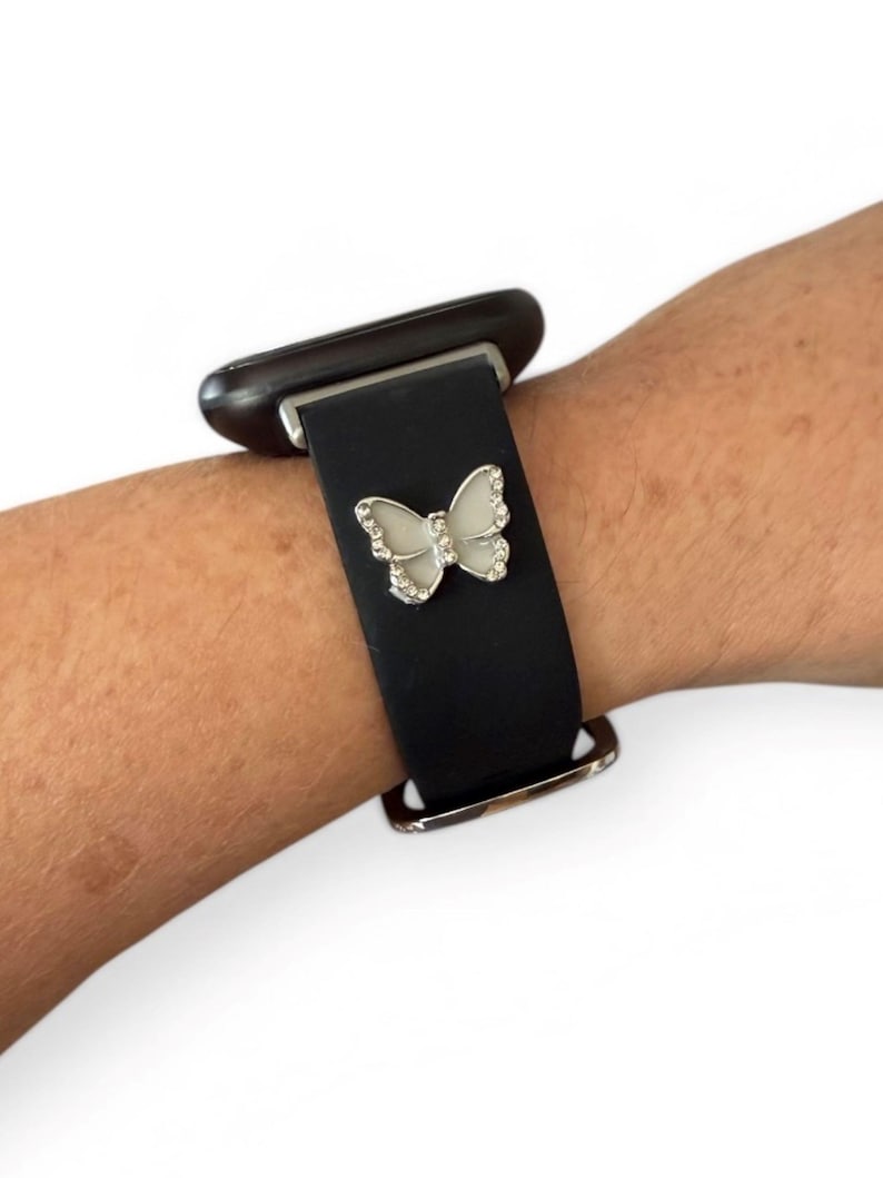 May include: Black watch band with a silver buckle and a decorative butterfly charm. The butterfly is white with silver-colored rhinestone accents. The watch band is attached to a black smartwatch.