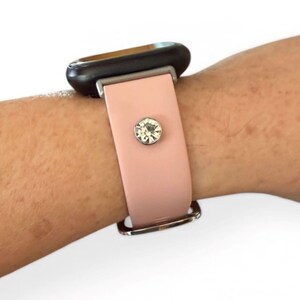 Peut inclure: Un bracelet de montre rose clair avec une boucle argent&eacute;e et un grand accent en cristal transparent. Le cadran de la montre est noir. Le bracelet est attach&eacute; &agrave; un poignet.