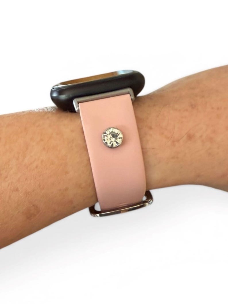 Peut inclure: Un bracelet de montre rose clair avec une boucle argent&eacute;e et un grand accent en cristal transparent. Le cadran de la montre est noir. Le bracelet est attach&eacute; &agrave; un poignet.