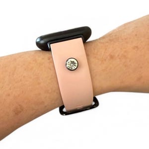 Peut inclure: Un bracelet de montre rose clair avec un bouton en cristal d&eacute;coratif. La montre est attach&eacute;e &agrave; un cadran noir. Le bracelet est port&eacute; sur un poignet.