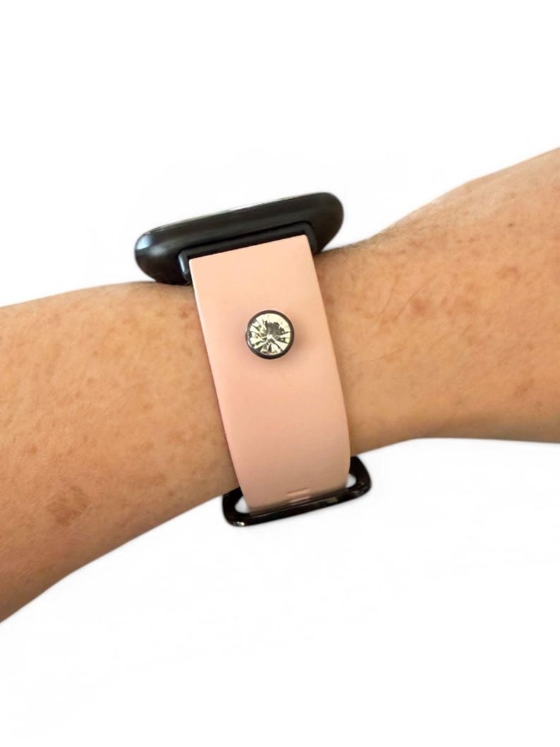 Peut inclure: Un bracelet de montre rose clair avec un bouton en cristal d&eacute;coratif. La montre est attach&eacute;e &agrave; un cadran noir. Le bracelet est port&eacute; sur un poignet.