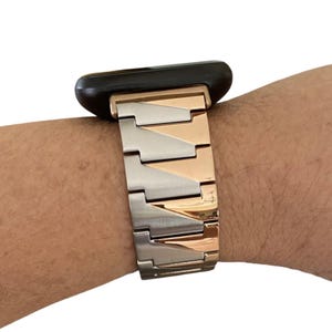 K&ouml;nnte beinhalten: Ein zweifarbiges Uhrenarmband in Silber und Ros&eacute;gold ist an einer schwarzen Smartwatch befestigt. Das Armband besteht aus rechteckigen Metallgliedern. Die Uhr befindet sich am Handgelenk einer Person.