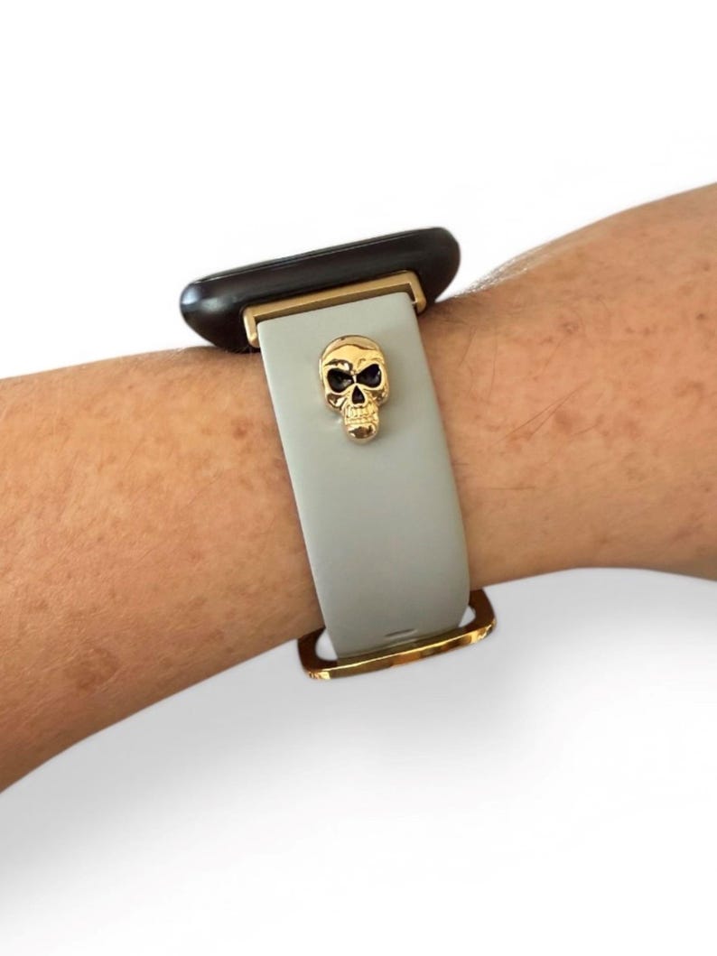 Op de afbeelding: Een lichtgrijze horlogeband met een gouden schedelbedel. De band heeft goudkleurige metalen accenten en is bevestigd aan een zwarte wijzerplaat. De schedelbedel is gedetailleerd met oogkassen en een neusholte.