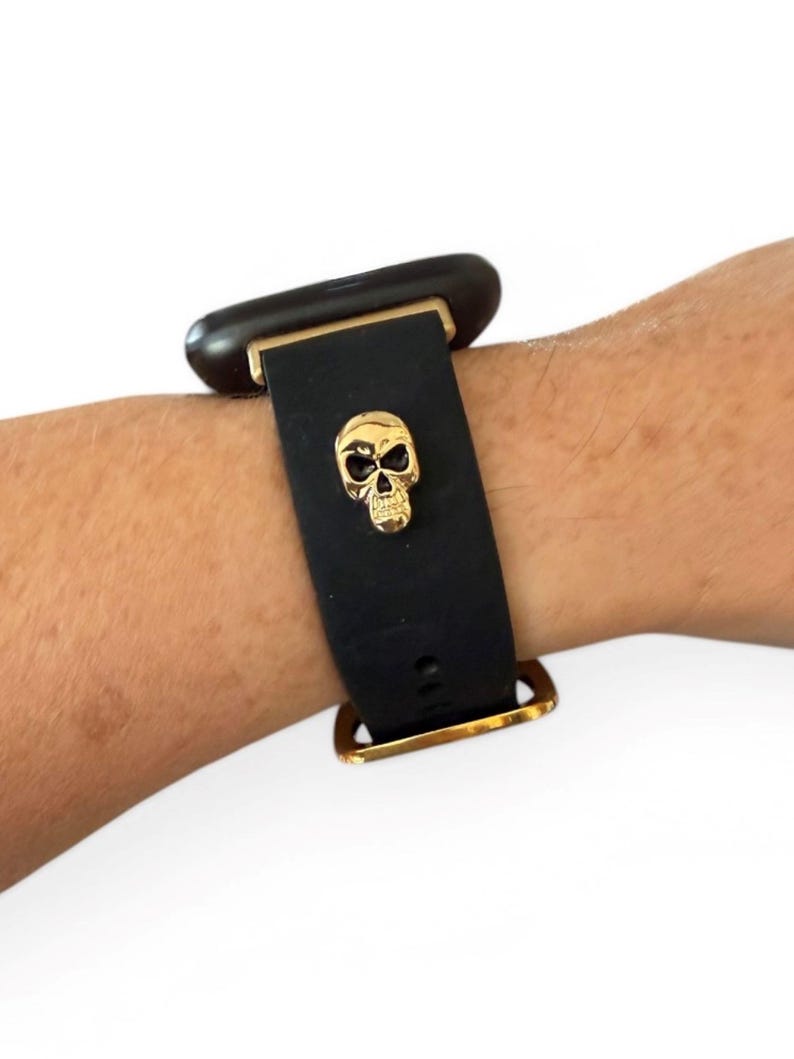 Op de afbeelding: Zwarte horlogeband met een gouden schedelbedel. De band is gemaakt van flexibel materiaal en heeft goudkleurige metalen hardware. De schedelbedel is gecentreerd op de band en voegt een unieke, edgy stijl toe.