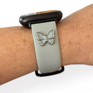May include: A light gray watch band with a silver butterfly charm. The butterfly is adorned with small, clear crystals. The watch face is black and gold.