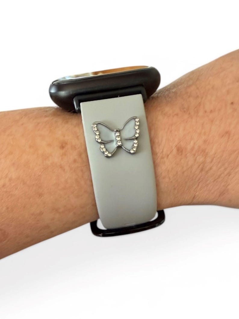 May include: A light gray watch band with a silver butterfly charm. The butterfly is adorned with small, clear crystals. The watch face is black and gold.