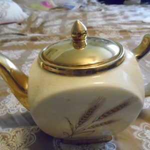 May include: A white teapot with a gold lid and handle. The teapot has a gold trim and a wheat design on the side.