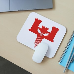 May include: White mousepad with a red castle and skull design. A white computer mouse sits below the mousepad. A blue spiral notebook and pencils are to the right. A laptop is in the background.