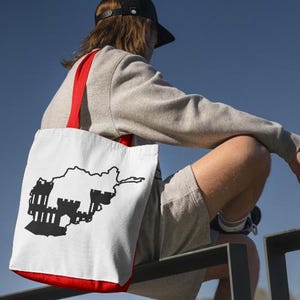 May include: White tote bag with a red trim and straps, featuring a black graphic of a country's outline and a castle. The bag is carried over the shoulder against a blue sky background.