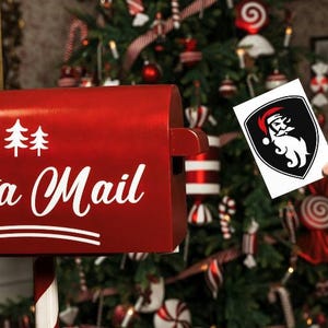 May include: A red Santa Mail mailbox with white text and a Christmas tree in the background. A person is holding a card with a Santa Claus graphic. The mailbox has a white stripe and two white Christmas trees.