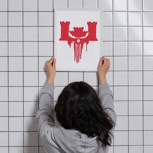 May include: A framed art print featuring a red graphic design of a castle-like structure with a skull and dripping elements. The artwork is held against a white tiled wall.
