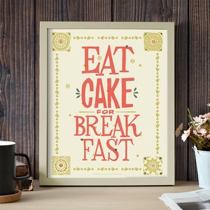 Może przedstawiać: Wydruk w ramie z białym tłem i złotymi akcentami. Wydruk zawiera tekst "Eat Cake for Breakfast." w czerwonych i złotych literach.