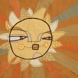 May include: A whimsical sun with a stitched face, brown eyes, and a stitched smile. The sun is made of fabric and has a yellow background with brown stitching. The sun is surrounded by orange and brown fabric rays.