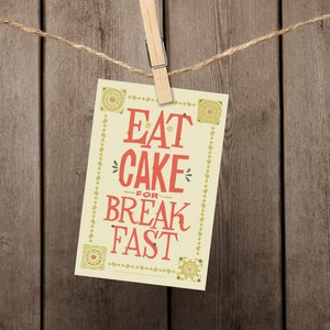 Może przedstawiać: Ż&oacute;łta kartka z ozdobną ramką wisi na sznurku z drewnianym klips. Na kartce widnieje napis "Eat Cake for Breakfast" czerwonymi literami.