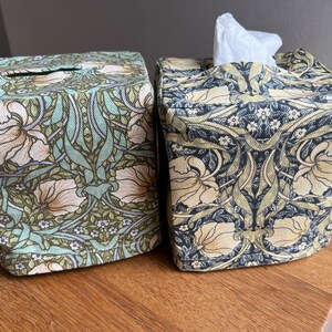 May include: Two square tissue box covers with a floral pattern. The cover on the left is green with white flowers, and the cover on the right is black with yellow flowers.