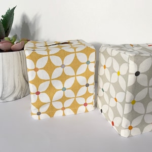 May include: Two tissue box covers, one yellow with white flowers and the other grey with white flowers. Both covers have colourful accents in the centre of the flowers.