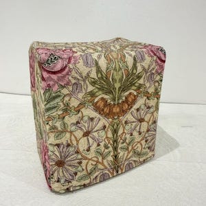 May include: A square ottoman with a floral pattern in pink, green, and purple on a beige background. The fabric design features stylized flowers and foliage, creating a vintage aesthetic. The ottoman is a cube shape.