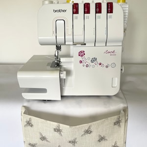 May include: A white Brother Lock M343D serger sewing machine with red and yellow details. The machine features a floral design and sits above a beige fabric catch bin with a grey bee pattern.